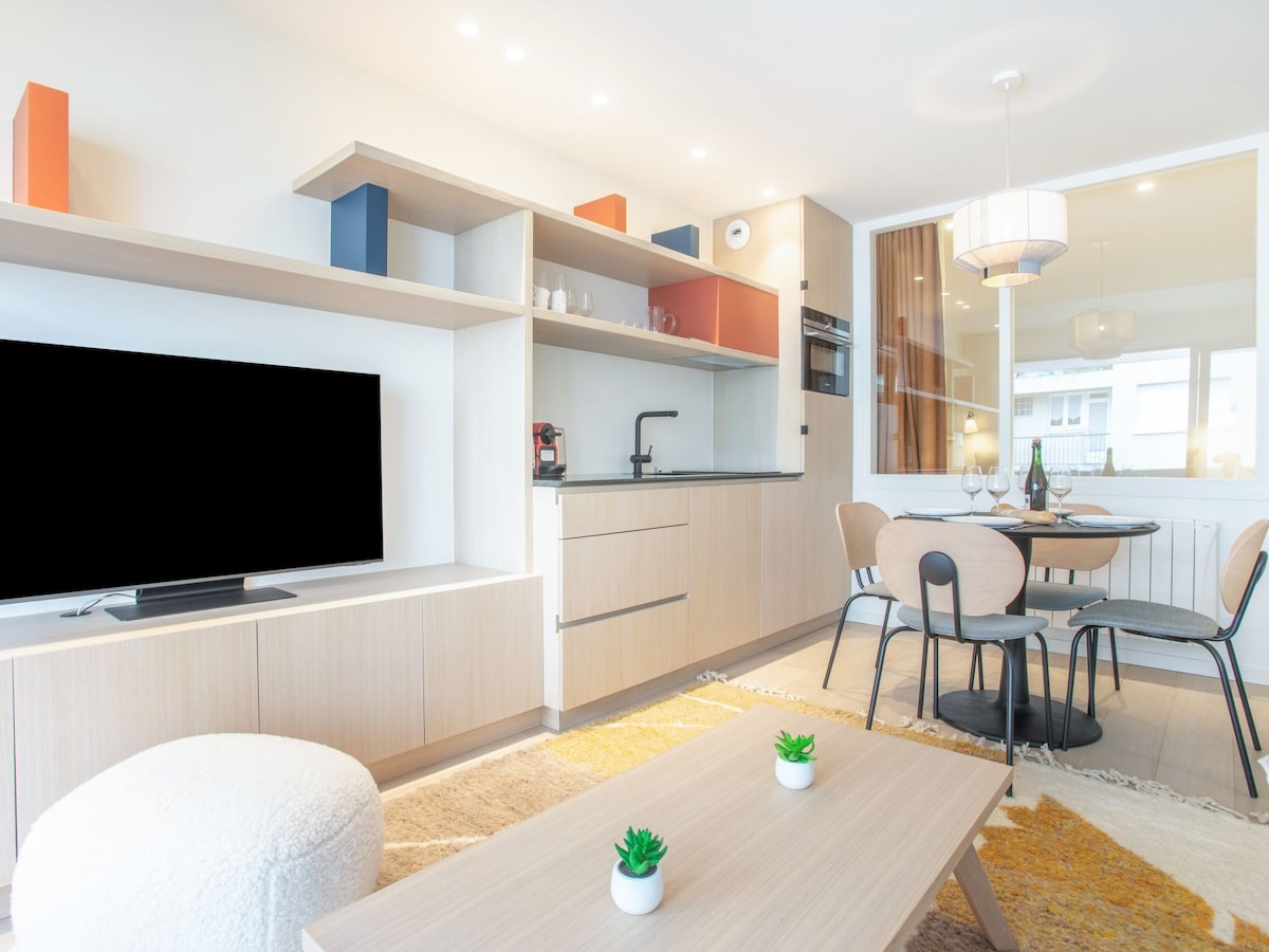 A modern living area features a sleek kitchenette with built-in appliances, including a coffee maker. A flat-screen TV is positioned in front of a minimalist coffee table, while a round dining table is set for two beside a large window, allowing ample natural light.
