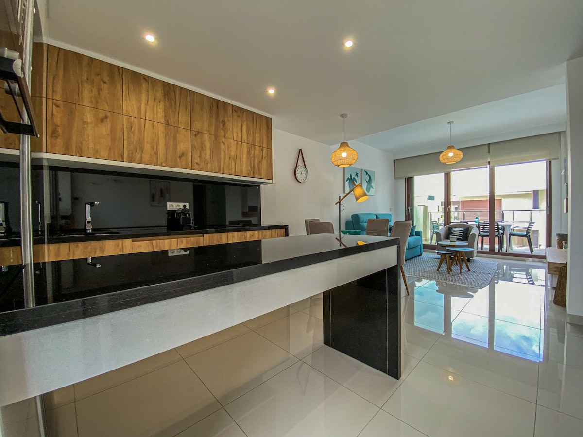 A modern kitchen area features wood cabinetry and a sleek black countertop, with a partial view of an open living space. Light filters in through sliding glass doors, illuminating the glossy tiled floors. Furnishings include comfortable seating and a dining table, enhancing the inviting layout.