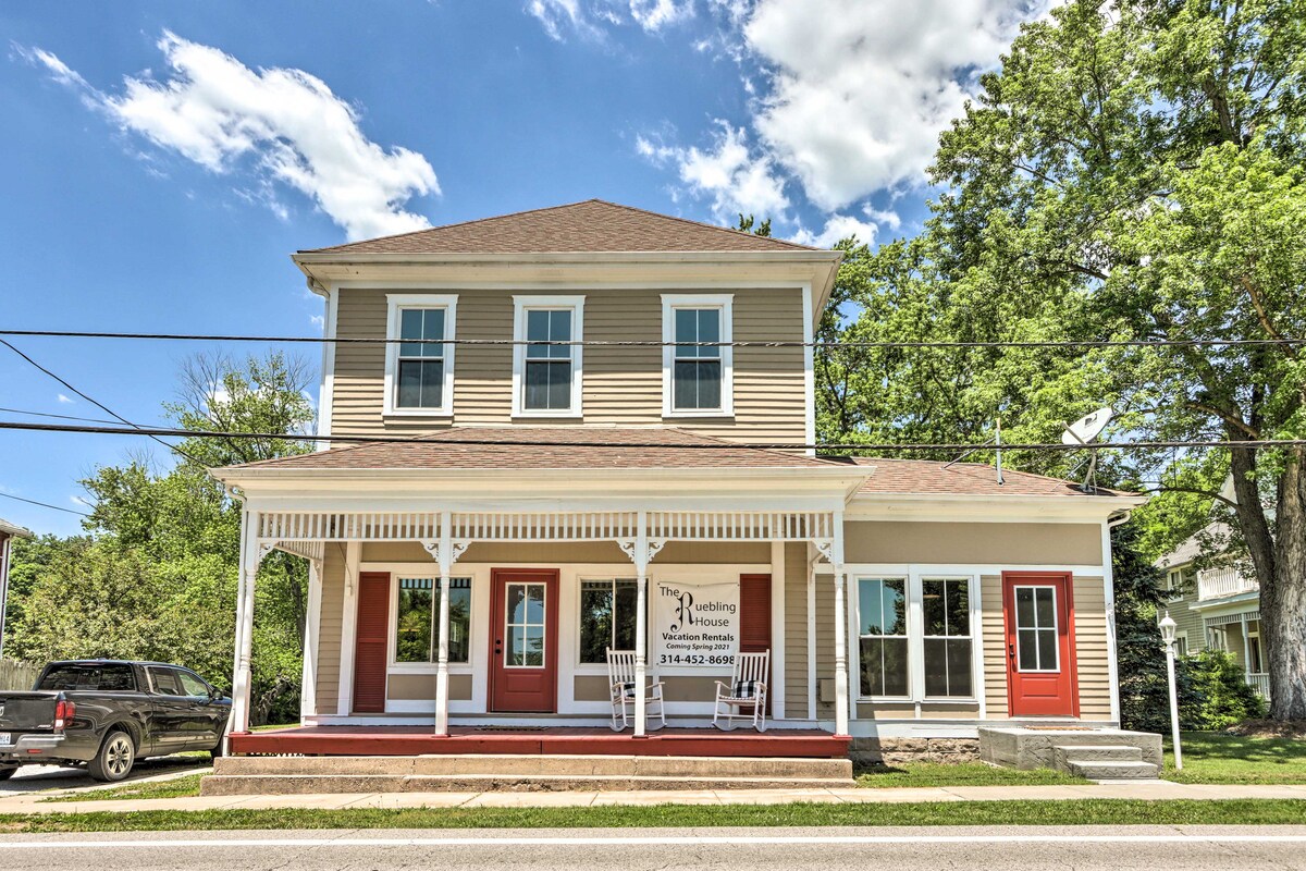 The exterior of the Ruebling House features a two-story structure with a beige facade and a red door. A covered porch with white railings is visible, providing a welcoming entryway. The surrounding greenery includes trees that enhance the rural charm.