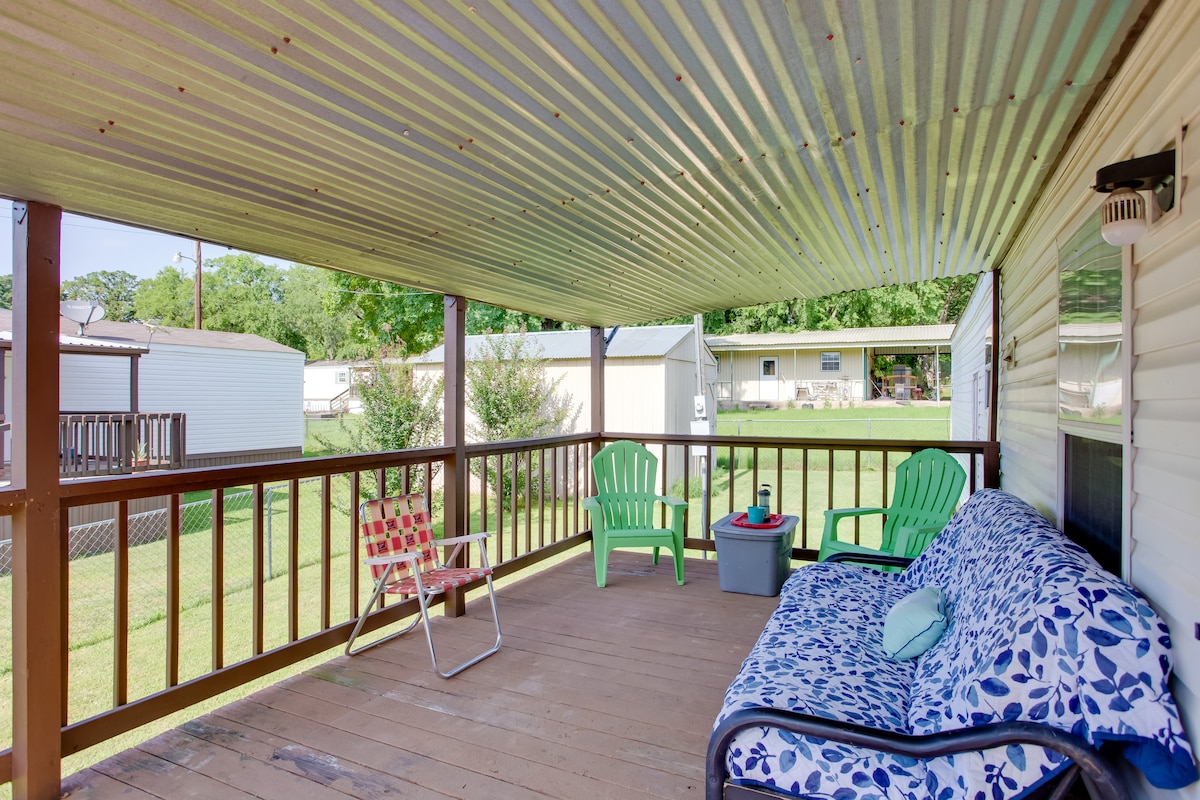 A covered deck is presented with a mix of seating options, including a patterned couch, a lounge chair, and two green adirondack chairs. The area is shaded by a corrugated metal roof, allowing for relaxation while overlooking a grassy yard.