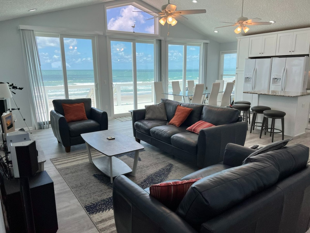 A spacious living area is furnished with dark leather sofas and a light coffee table, all oriented towards large windows showcasing ocean views. Natural light floods the room, enhancing the airy feel. A dining area with white chairs and a kitchen with dual refrigerators are visible in the background.