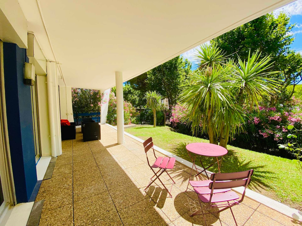 A spacious terrace is surrounded by lush greenery, featuring a pink round table and two matching chairs. Sunlight filters through the open space, illuminating the decorative plants that enhance the outdoor area, creating a relaxing environment.