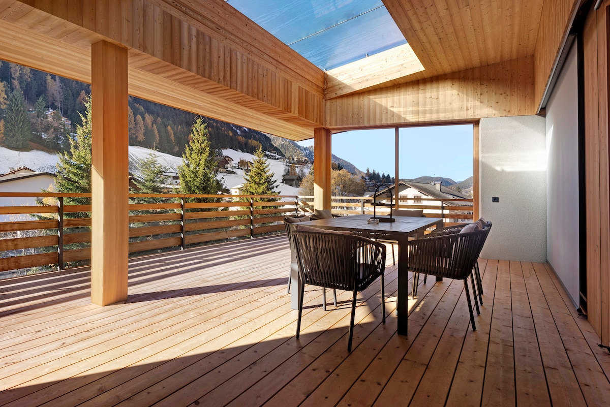 An outdoor terrace features a spacious wooden dining table surrounded by elegant chairs. Large windows provide a panoramic view of the surrounding mountains. The ceiling is partially transparent, allowing natural light to illuminate the area, enhancing the connection with the outdoors.