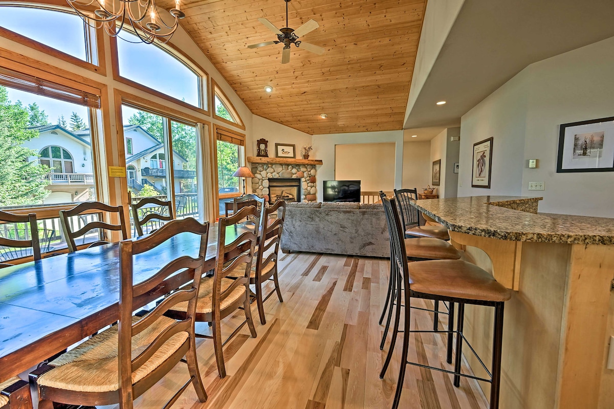 A spacious dining area features a large wooden table surrounded by several wooden chairs, while a counter with bar stools offers additional seating. Large windows fill the space with natural light, showcasing views of the surrounding greenery and inviting a relaxed ambiance.