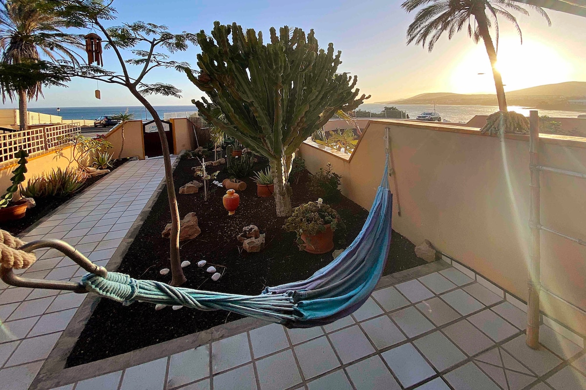 A serene outdoor space is depicted, featuring a colorful hammock suspended between tree branches. Lush plants and decorative pots populate the area, while a vibrant sunset casts a warm glow over the garden. The ocean is visible in the background, enhancing the tranquil ambiance.