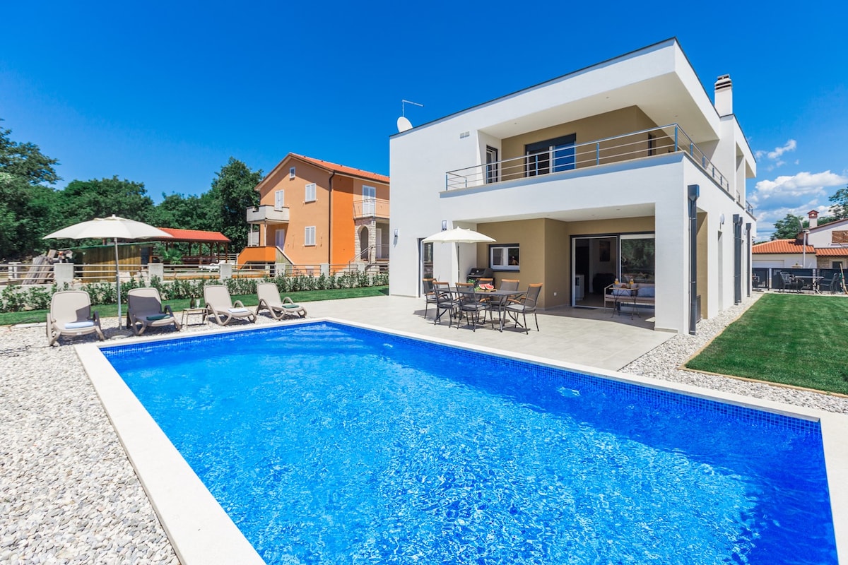 A modern home features a private swimming pool with clear blue water surrounded by sun loungers. An outdoor dining area is visible, offering a comfortable setting for meals. The landscaped garden includes green space and decorative stones, contributing to a serene outdoor environment.