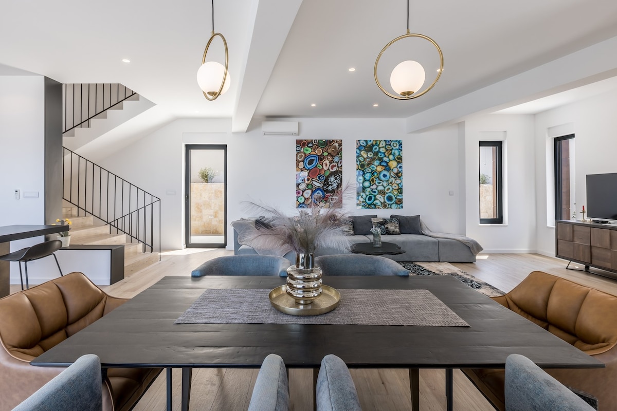 An open-concept living and dining area features a large black dining table surrounded by plush chairs. Colorful wall art adds visual interest, while a sectional sofa offers ample seating. Natural light filters through large windows, illuminating the space.