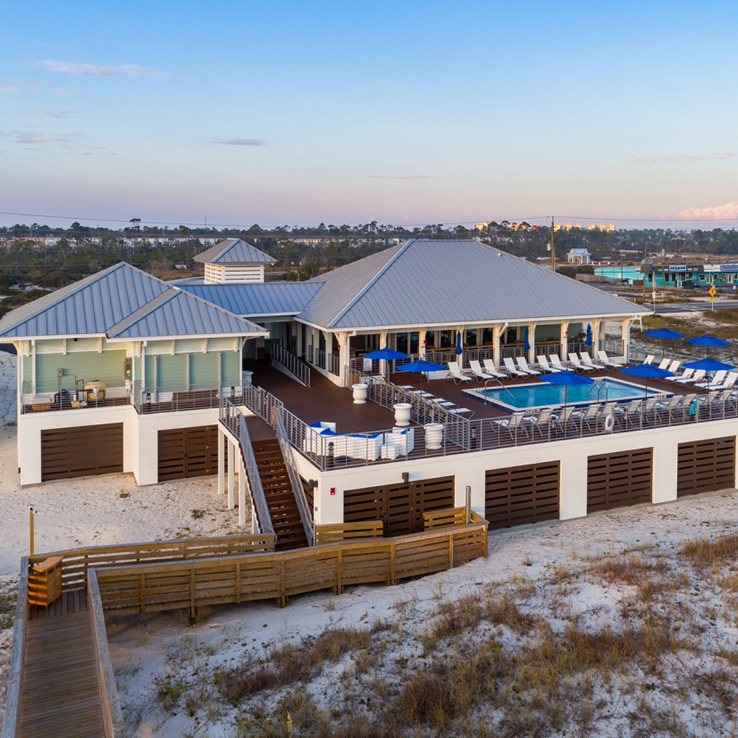 The beach club facility features a spacious deck with multiple lounge chairs surrounding a large swimming pool. The modern structure has a sloped metal roof and large windows, providing views of the surrounding area. The sandy pathway leads directly to the beach.