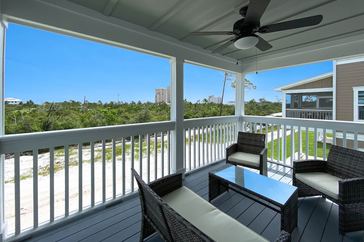 A covered balcony is furnished with a seating arrangement, including two chairs and a coffee table. Lush green landscapes and distant beach views are visible beyond the railing, while bright blue skies provide a cheerful backdrop.