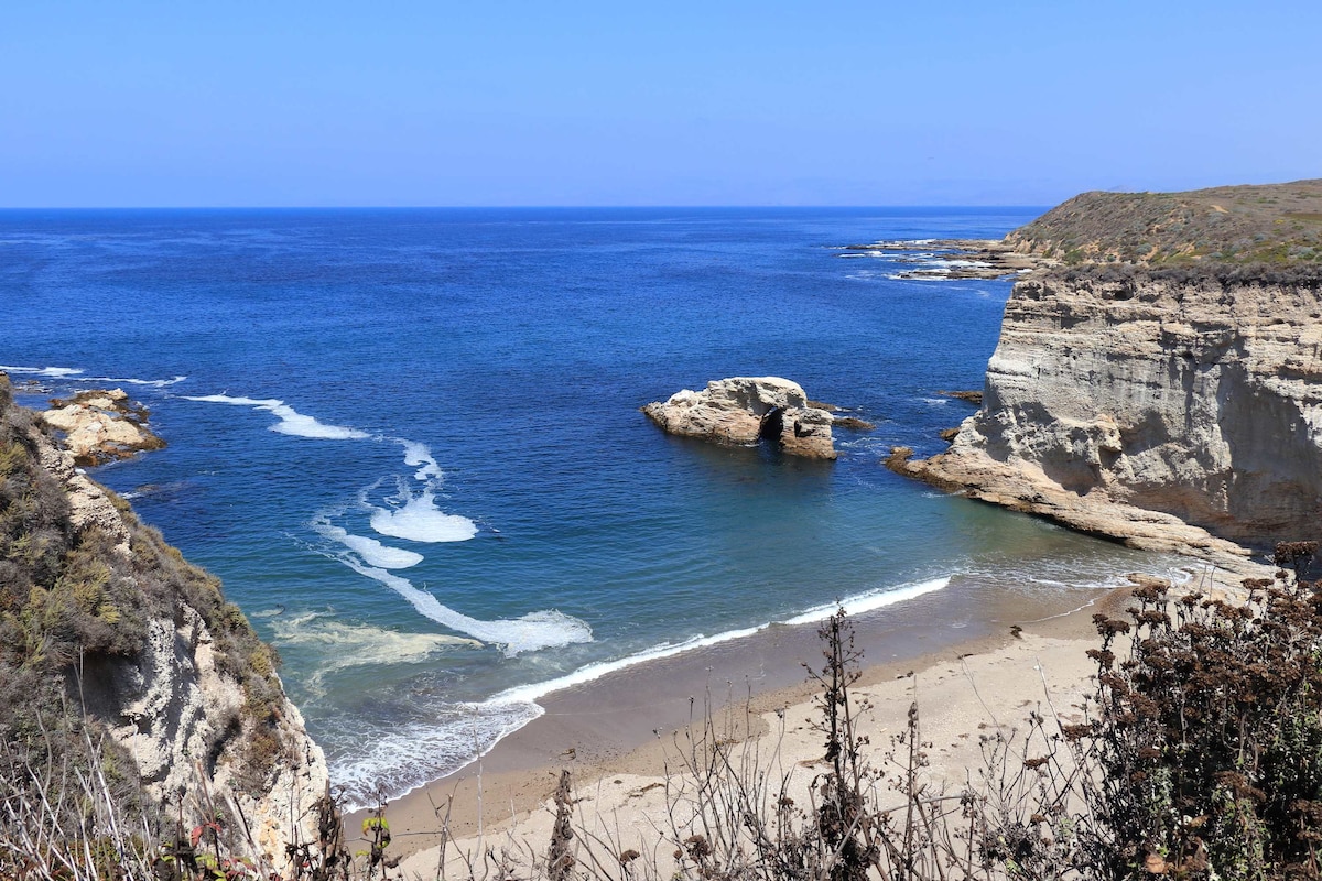 A serene coastal view showcases a sandy beach bordered by rocky cliffs. Gentle waves lap against the shore, with clear blue waters extending into the ocean. A calm, sunny atmosphere is present, enhanced by the vibrant blue sky above.