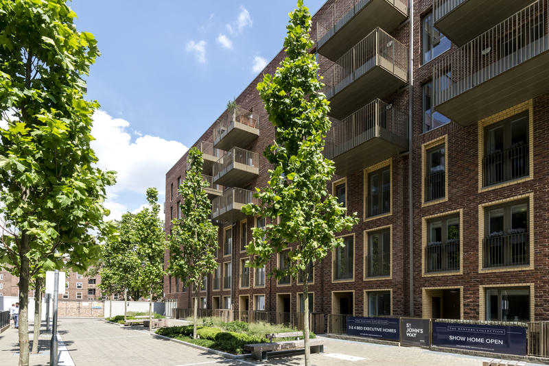 The exterior of a modern apartment building is shown, featuring a brick facade and multiple balconies. Lush trees line the pathway in front, providing greenery. Clear blue skies are visible overhead, enhancing the tranquil setting of the complex.