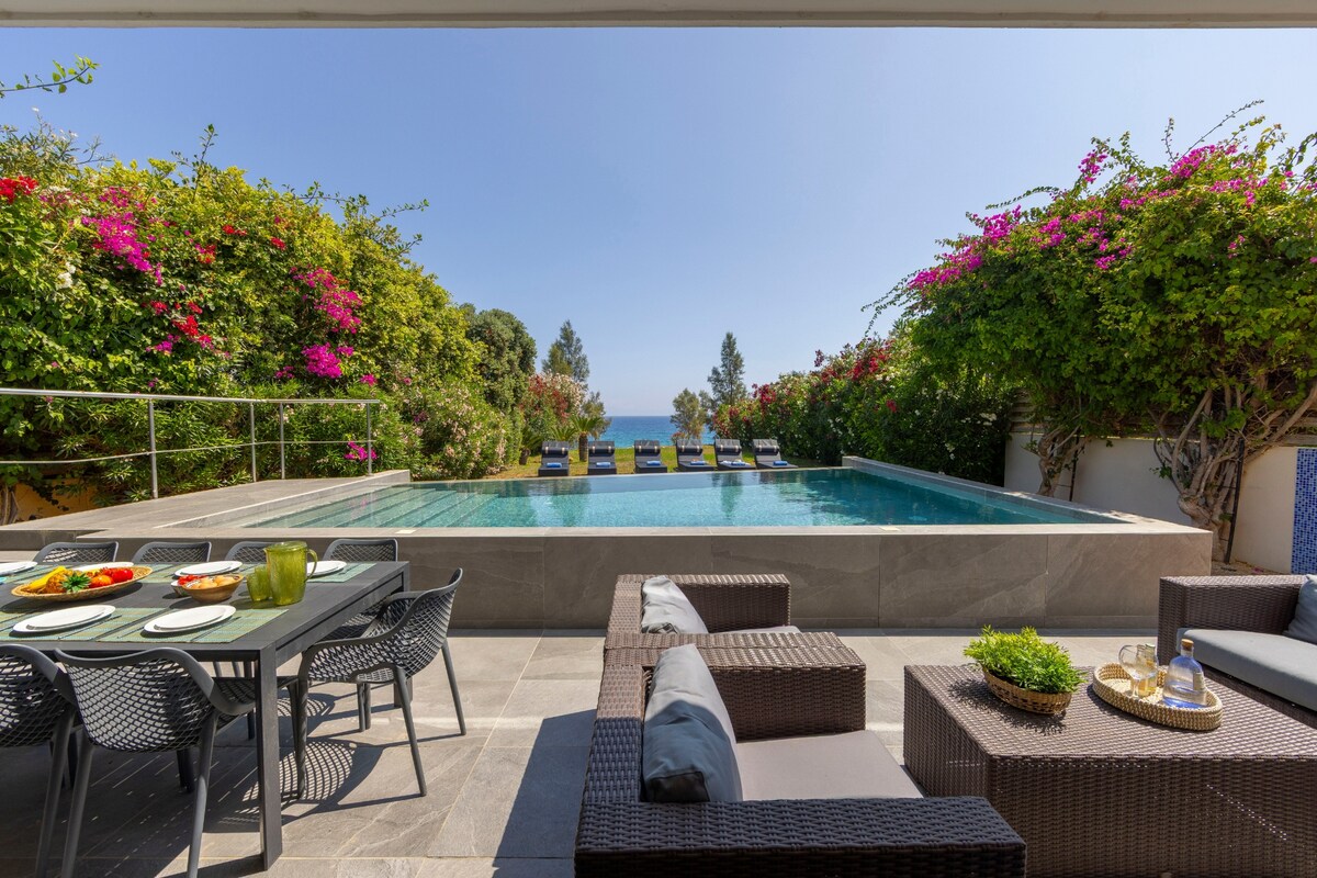 An outdoor space features a private pool surrounded by lush greenery, with colorful flowering plants bordering the area. A modern seating arrangement is complemented by a dining table set for outdoor meals, overlooking the tranquil pool and garden, with distant sea views.