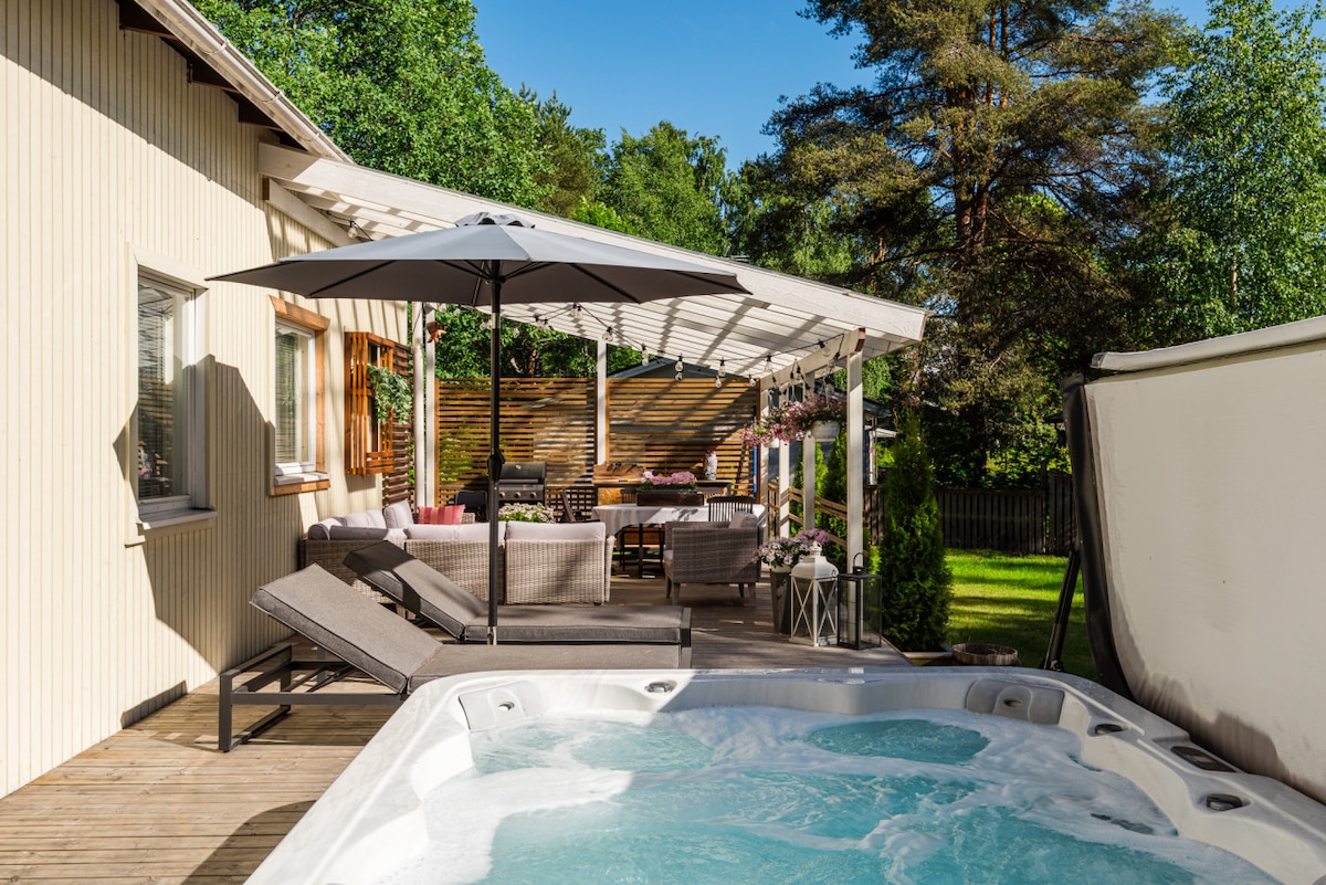 An outdoor terrace is featured, complete with comfortable lounge chairs and a shaded dining area. A hot tub invites relaxation, surrounded by greenery and sunlight casting a warm glow over the space. The area provides a perfect setting for outdoor gatherings.