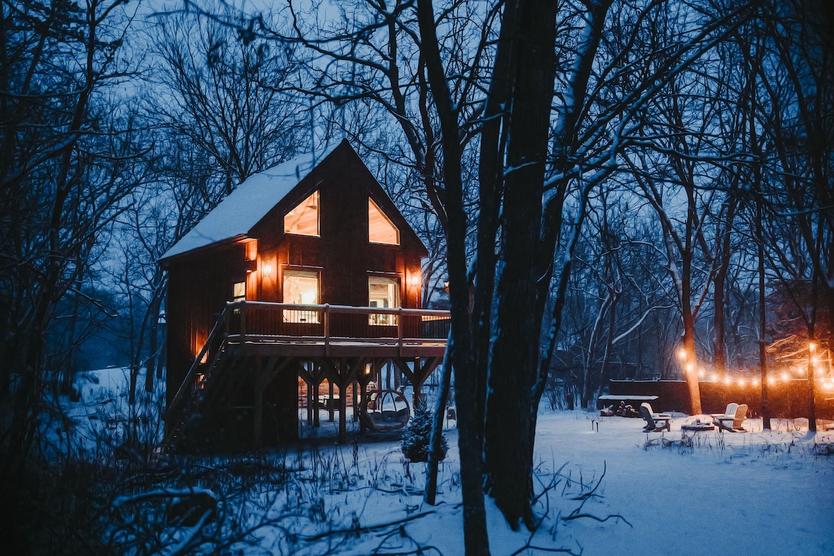 The treehouse stands amidst a snowy landscape, highlighted by warm lights emanating from its windows. The surrounding trees are bare, complementing the tranquil evening atmosphere. An outdoor seating area with string lights creates a cozy ambiance, inviting relaxation in the serene woodland setting.