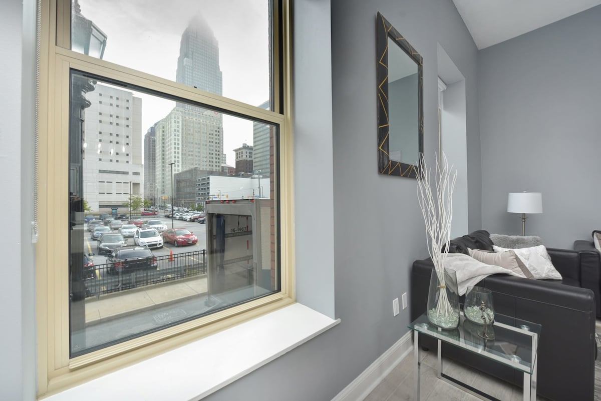 A large window offers a view of the city skyline and a nearby parking area. Natural light fills the room, highlighting the gray walls and modern decor. A stylish mirror and decorative vase on a glass table complement the cozy seating area.