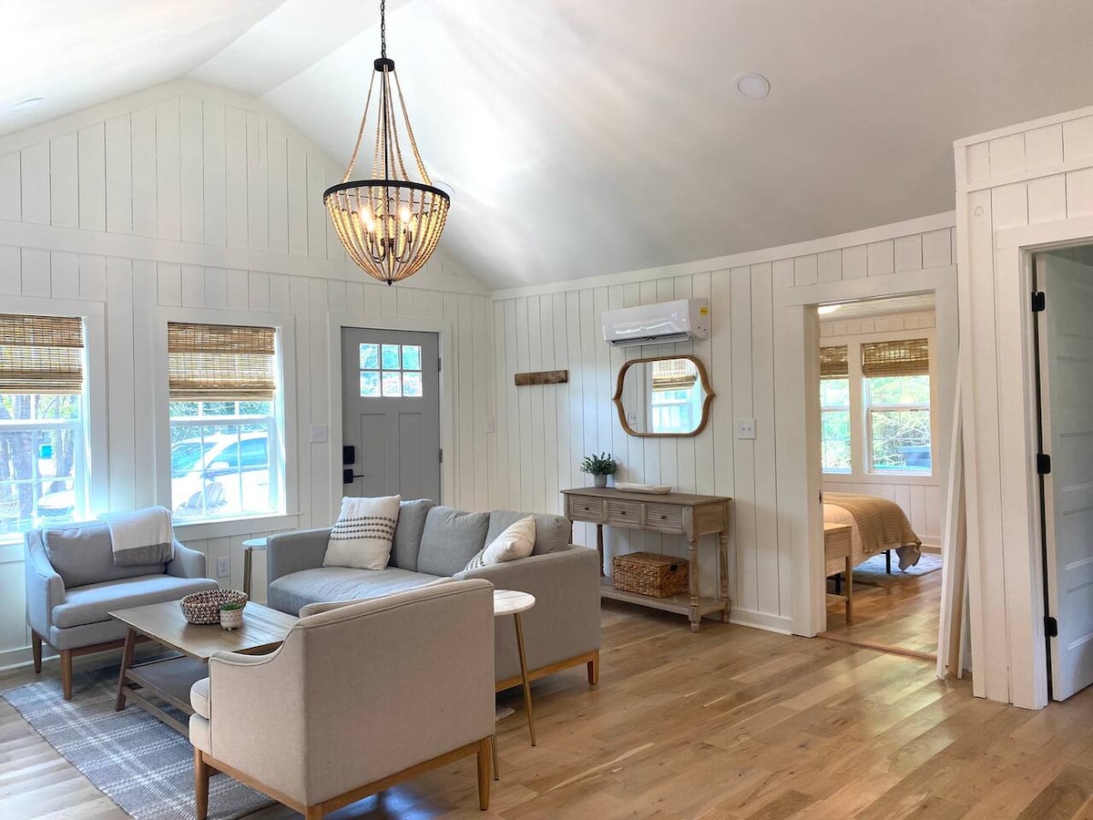 A spacious living area featuring vaulted ceilings and a stylish chandelier. Light-colored shiplap walls enhance the room's brightness, while large windows allow natural light to fill the space. Contemporary furniture includes a cozy sofa, a wooden coffee table, and a buffet table, providing both comfort and functionality.
