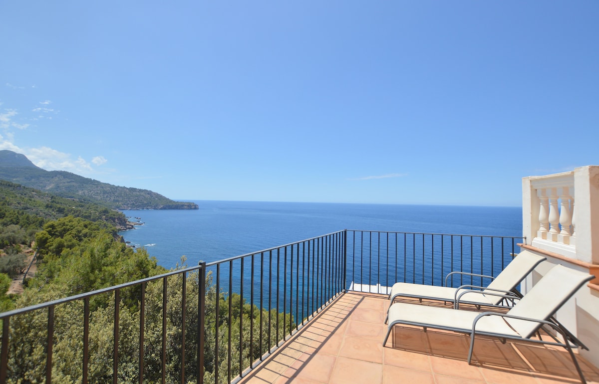 A sun terrace is featured with two lounge chairs positioned for optimal views of the Mediterranean Sea. The surrounding landscape includes lush greenery and mountainous terrain, with a clear blue sky enhancing the coastal scenery.