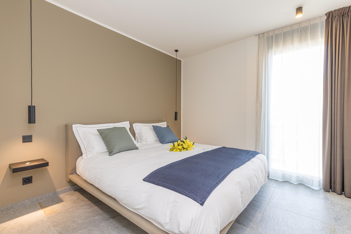 A spacious bedroom features a large double bed adorned with decorative cushions. Beside the bed, minimalist wall-mounted lamps provide soft lighting. Natural light enters through a large window, partially covered by elegant sheer curtains, highlighting the modern design of the room.