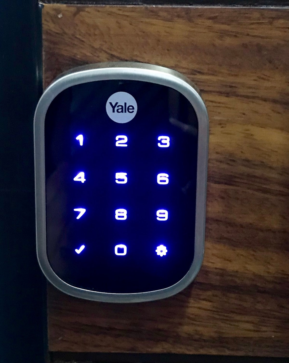 A keyless entry pad is prominently displayed, featuring illuminated numbers from 0 to 9. The secure lock, designed by Yale, is integrated into a wooden surface, providing a modern touch. The sleek design is user-friendly, ensuring easy access to the property.