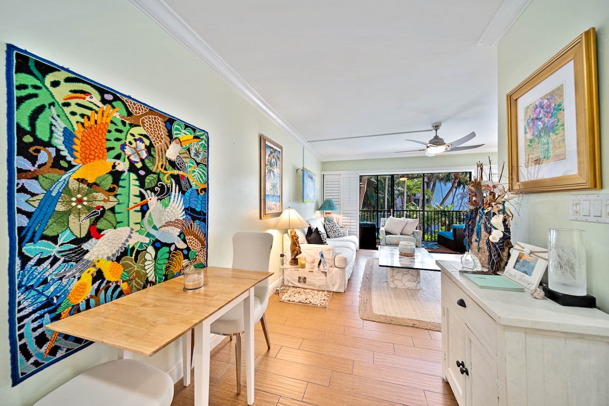 The living area is characterized by a vibrant wall tapestry depicting tropical birds, complemented by light-colored furnishings. A wooden dining table is positioned near the entrance, while large windows provide a view of the outdoor space filled with greenery.