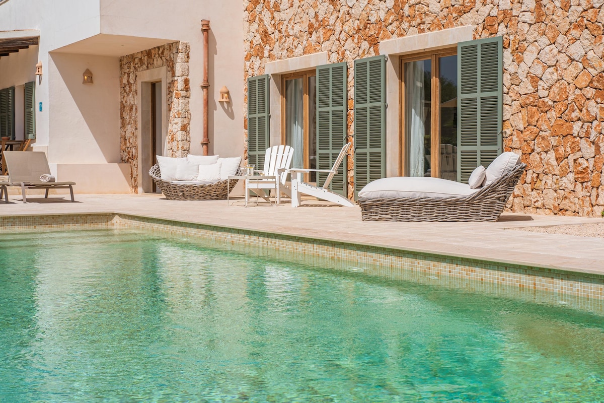 A serene outdoor area features a clear swimming pool, bordered by a stone wall and comfortable lounge chairs. Large windows with green shutters are visible, allowing natural light to enhance the space. Textured furniture complements the poolside setting.