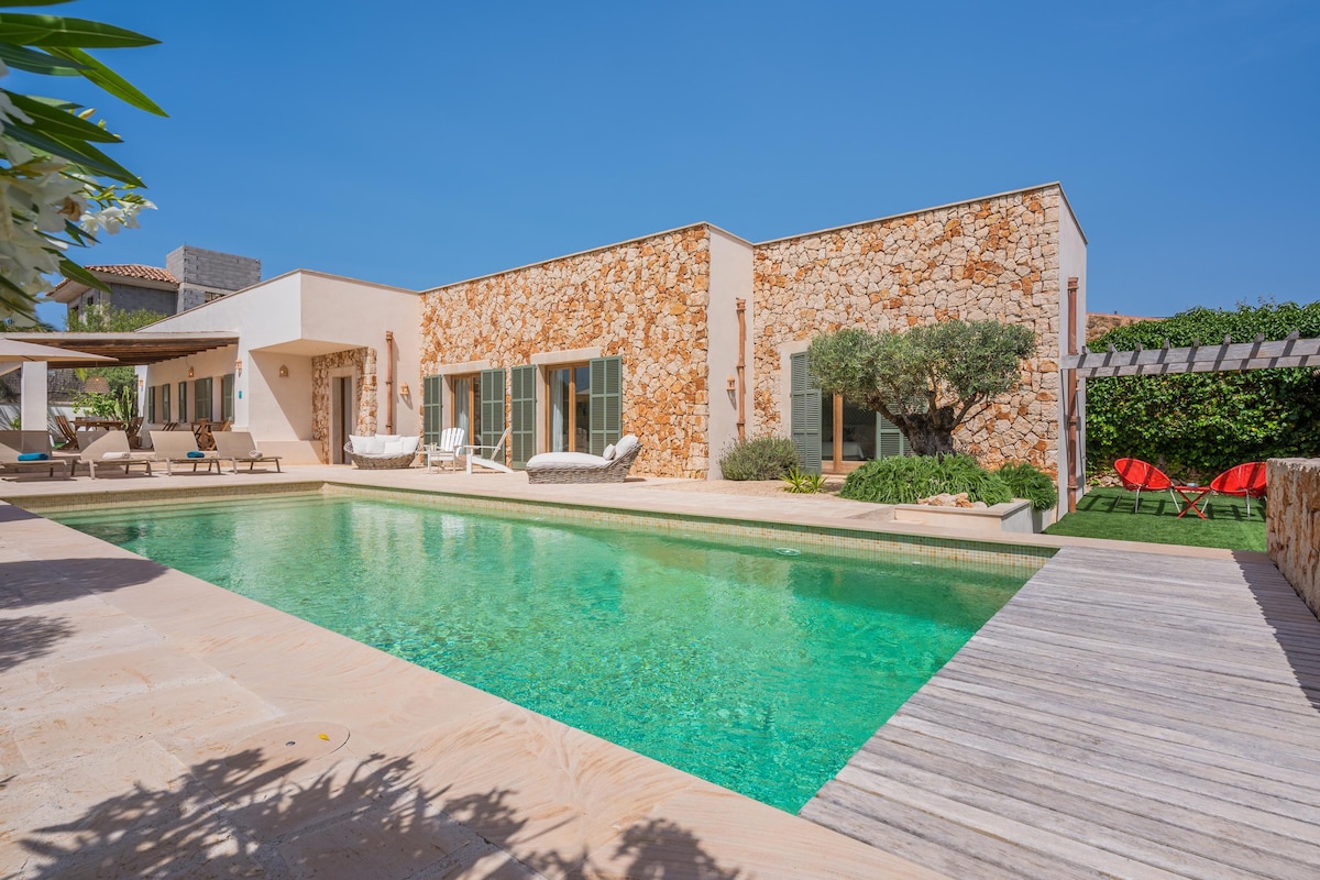 A modern villa with a natural stone facade is surrounded by lush greenery and features a sparkling pool. Comfortable loungers and bright red chairs are positioned by the water, creating an inviting outdoor space for relaxation under the clear blue sky.