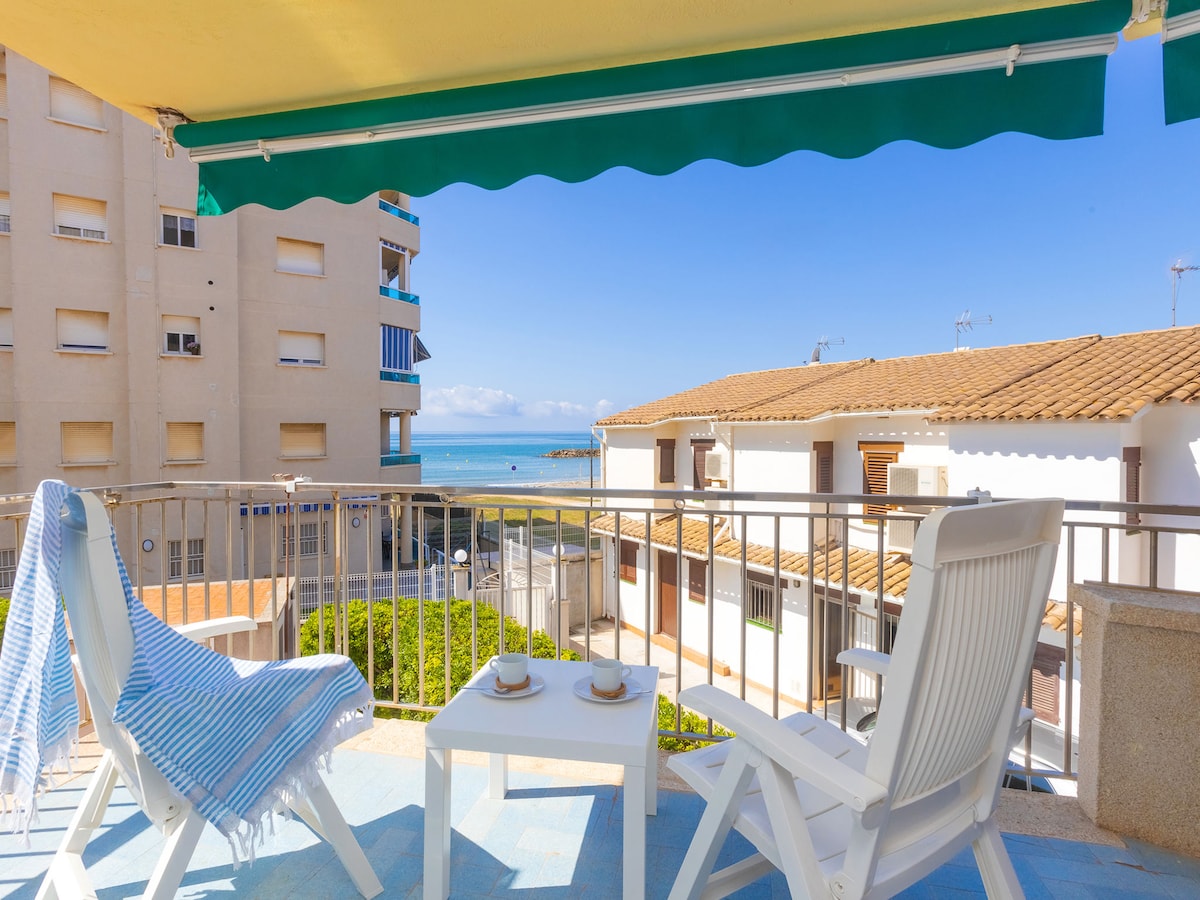 A balcony with two white chairs is shown, adorned with a blue and white striped throw blanket. A view of the sea is visible in the background, complemented by clear blue skies and distant buildings. Small cups are positioned on the table between the chairs.