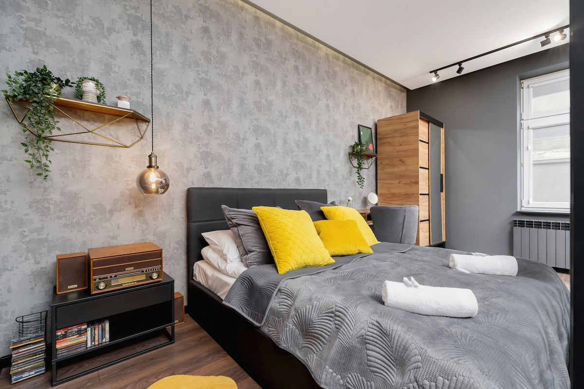 Udobna spavaća soba s tapetama, uredno namješten krevet sa sivom posteljinom i žutim ukrasnim jastucima. U kutu se vide radni stol i police.