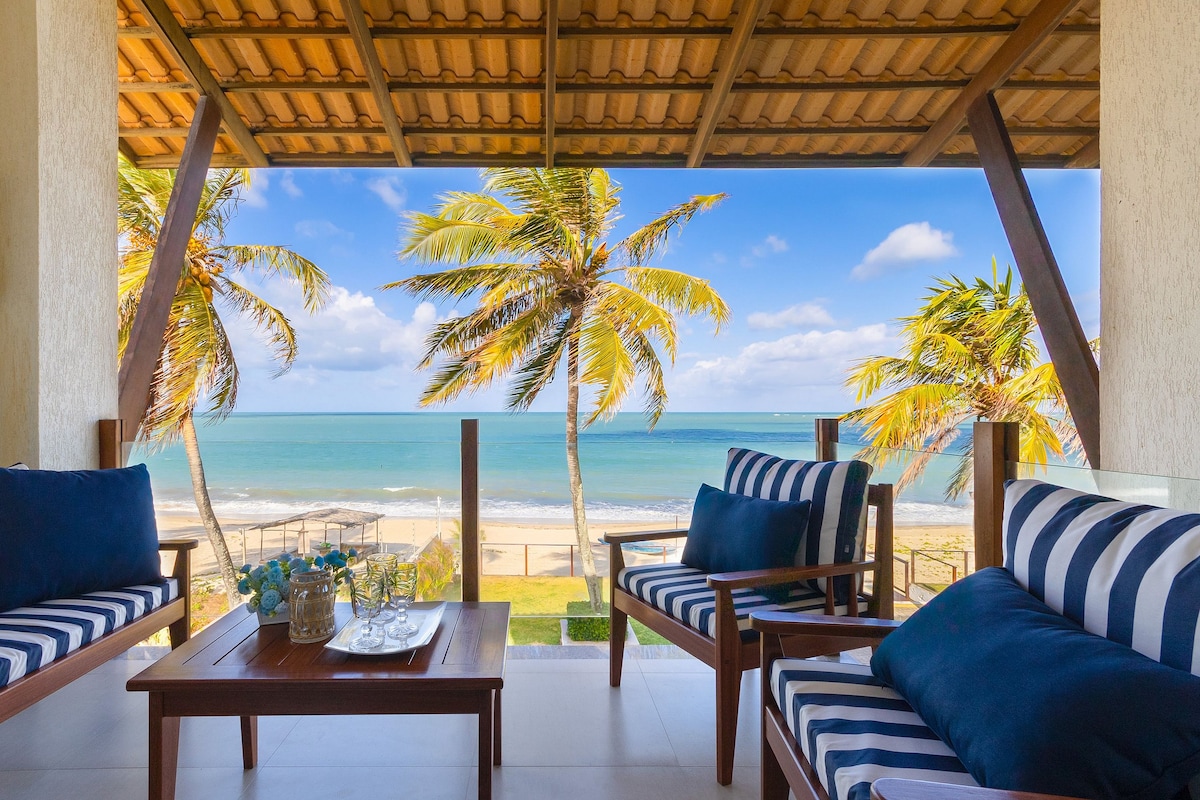 A covered terrace offers a view of the beach and ocean, framed by swaying palm trees. Comfortable seating arranged around a wooden table invites relaxation, while blue and white striped cushions provide a coastal touch. The space is filled with natural light.