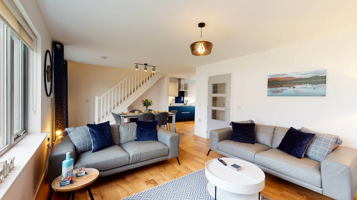 A light-filled open-plan living area is shown, featuring two comfortable sofas facing a central coffee table. A stylish light fixture hangs overhead, and a dining space with blue cabinetry is visible in the background. Locally inspired artwork adorns one wall, enhancing the welcoming ambiance.