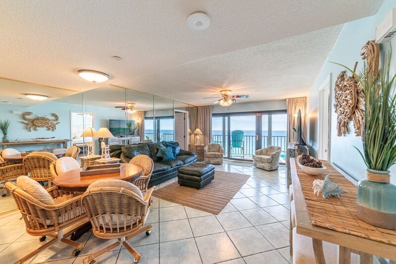 A spacious living area is highlighted by large windows that provide direct views of the beach. Comfortable seating is arranged around a coffee table, complemented by a dining table surrounded by wicker chairs. The decor features neutral tones, enhancing the inviting atmosphere.