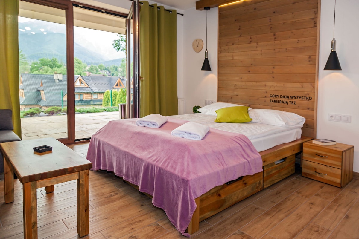 A cozy bedroom features a spacious bed with a light purple blanket and neatly folded towels on top. Wooden accents are highlighted on the walls and furniture, while large windows offer natural light and views of the surrounding mountains.