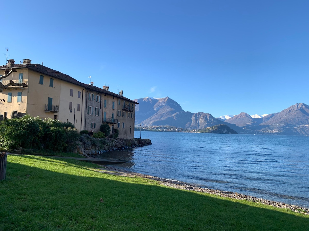 A lakeside view is presented, featuring a charming building with multiple windows and balconies. The calm waters of Lake Como reflect the surrounding mountains under a clear blue sky. Lush greenery and a pebbled shore can be seen in the foreground.