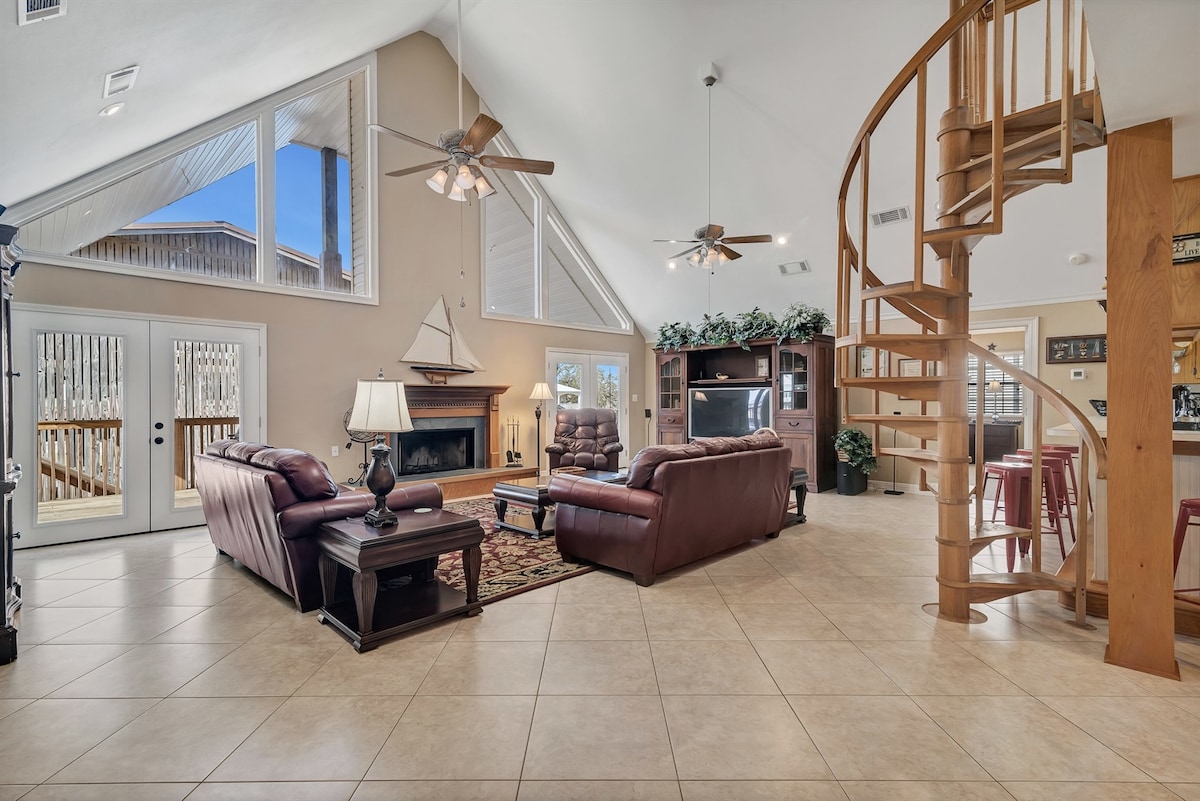 The spacious living room features high cathedral ceilings and tiled flooring, accented by a cozy fireplace. Comfortable leather seating is arranged for relaxation, with a large flat-screen TV visible. A spiral staircase leads to the upper loft area, enhancing the home's inviting ambiance.