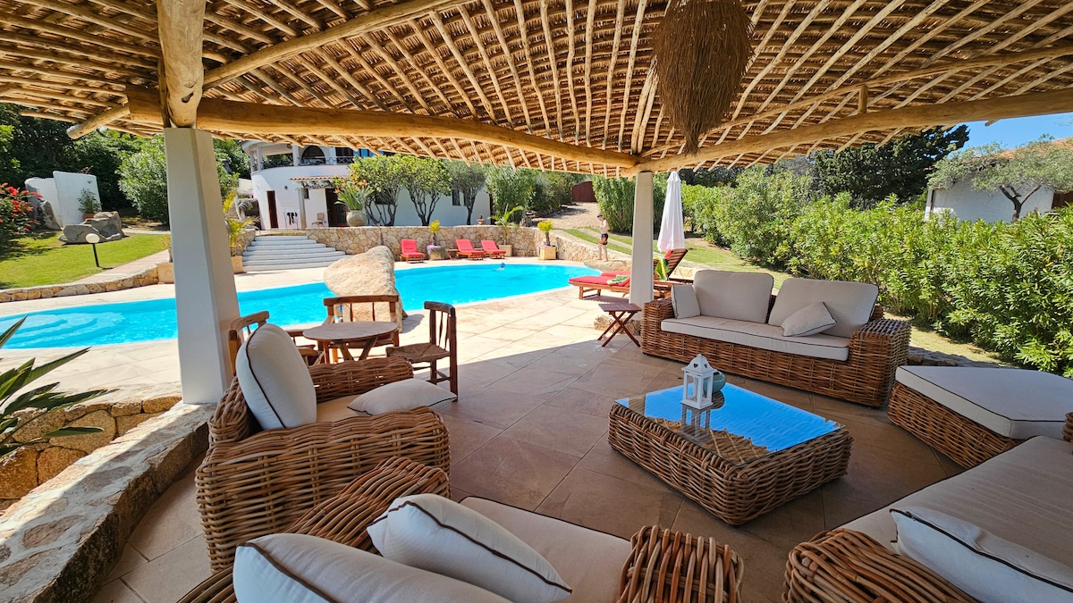 A shaded seating area features comfortable wicker furniture arranged around a glass-top table. The space provides a view of the inviting blue pool surrounded by greenery and lounge chairs placed on the tiled terrace. The natural canopy above adds a rustic touch.
