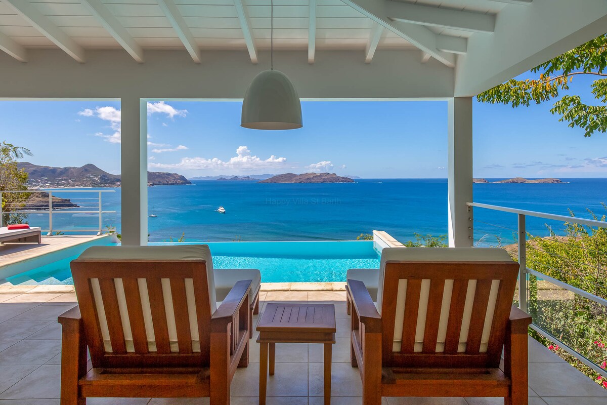 A serene view is offered from the terrace, showcasing a calm sea against a clear blue sky. Two wooden lounge chairs and a small table are positioned to enhance the outdoor lounging experience, while an infinity pool stretches out towards the horizon.