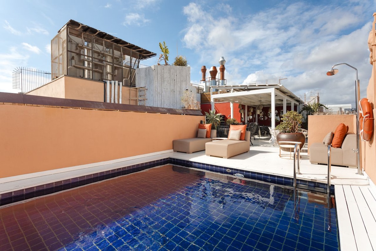 A rooftop terrace features a serene pool set against a bright sky. Comfortable lounge seating is positioned beside the water, surrounded by potted plants. A shaded dining area can be seen in the background, offering an inviting atmosphere for relaxation.
