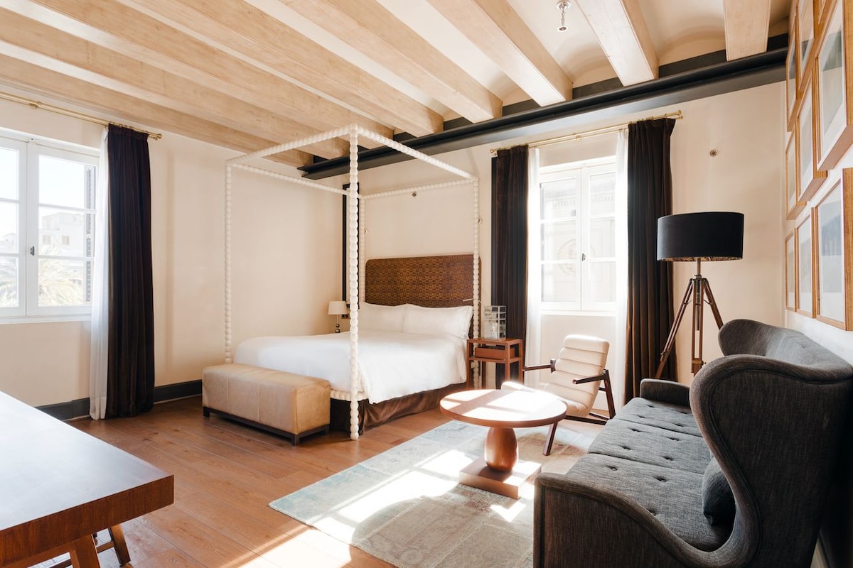 A spacious room features a four-poster bed with a woven headboard, complemented by a classic armchair and a modern side table. Natural light enters through large windows, while dark curtains frame the view. Wooden beams line the ceiling, enhancing the room's elegant design.