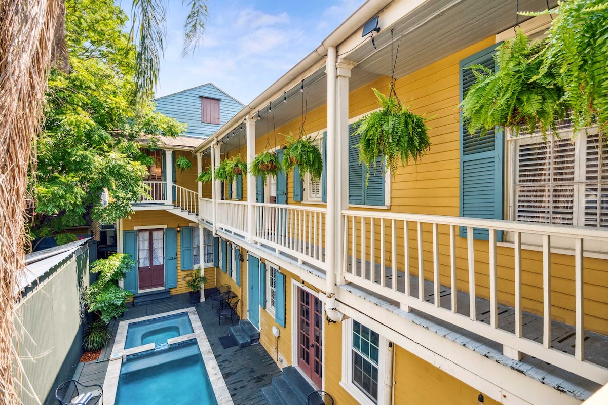 The exterior view of the restored mansion showcases a vibrant yellow facade with blue shutters. Lush greenery hangs from porches, while a serene heated pool is situated in the courtyard, surrounded by stone tiles and outdoor seating. Natural light brightens this inviting outdoor space.