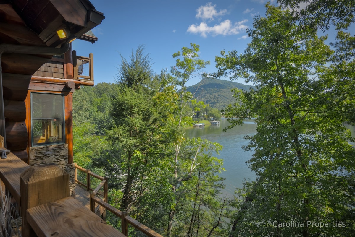 A scenic view is captured from the balcony of the log home, showcasing lush trees and a tranquil lake. The surrounding mountains rise in the background, enhancing the natural beauty of the area under a clear blue sky.