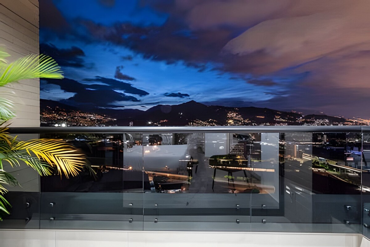 A private balcony with a glass railing offers a scenic view of the city skyline at dusk. The sky transitions from deep blue to light, with a glimpse of mountain silhouettes illuminated by city lights below, creating a serene outdoor setting.