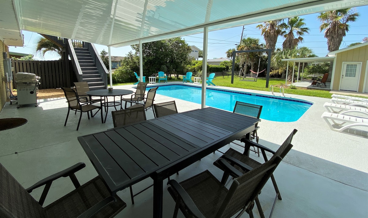 An outdoor living area features a large table surrounded by several chairs, providing ample seating. A turquoise pool reflects sunlight in the background, with lounge chairs arranged nearby. Palm trees and grassy space enhance the inviting environment, accessed via a staircase leading to a second level.