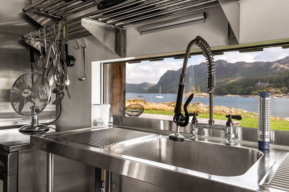 A modern kitchen space is showcased with a stainless steel sink and a large window framing a view of Loch Carron and the surrounding hills. An array of utensils can be seen hanging above the sink, contributing to a well-equipped cooking area.