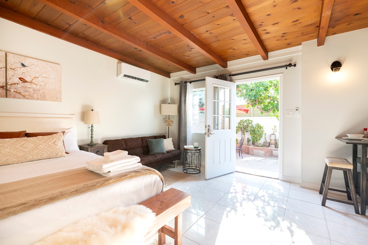 A well-lit studio space features a queen-sized bed with bedding and towels neatly arranged. A comfortable sofa and a small dining table are present. A set of double doors opens to the outdoor area, surrounded by greenery, inviting natural light into the room.