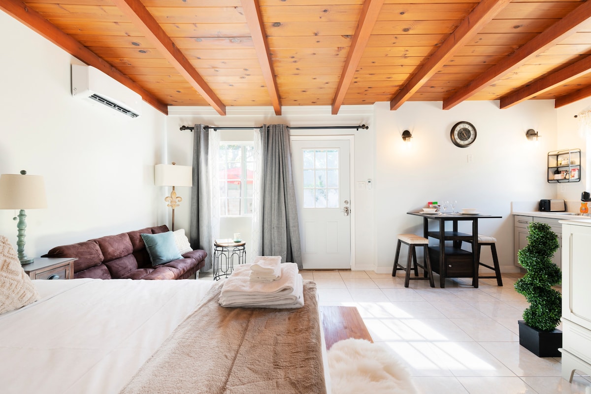 The studio interior showcases wooden beams across the ceiling, enhancing the spacious feel. A cozy sofa complements a dining table for two, while natural light streams through the front door. Fresh towels are neatly arranged on the bed, offering a sense of comfort.