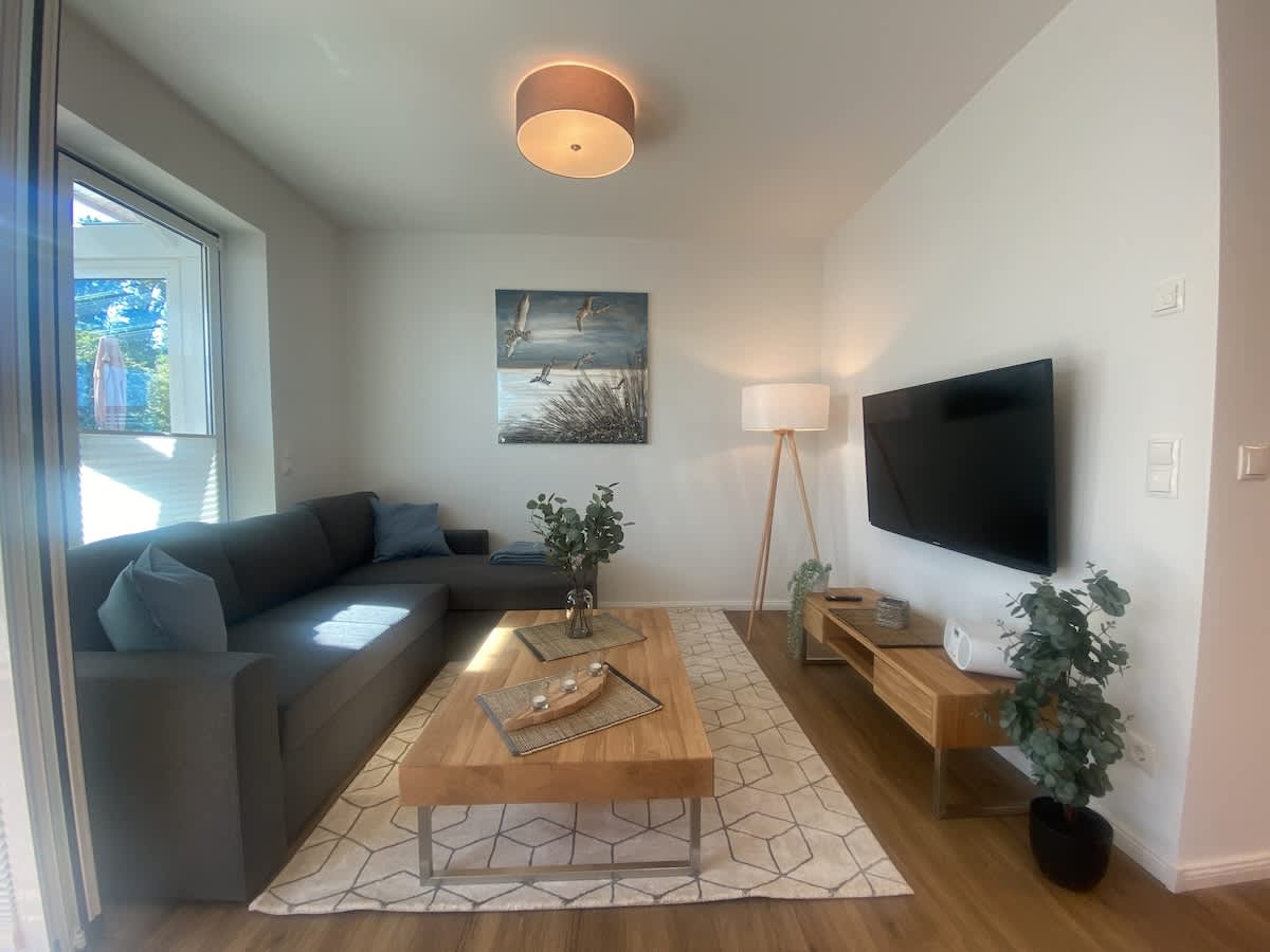 A modern living area showcases a spacious grey sectional sofa paired with a wooden coffee table. A flat-screen TV is mounted on the wall opposite the seating. Natural light pours in through a large window, highlighting the artwork above the sofa depicting a serene outdoor scene.