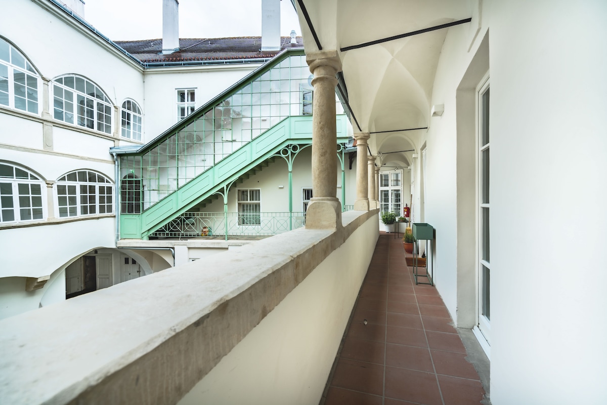 A spacious corridor features a long, light-colored railing with decorative details, overlooking a central courtyard. Greenery is visible on the balcony outside, along with large windows that provide natural light. The tiled flooring and archways add to the historical charm of the space.