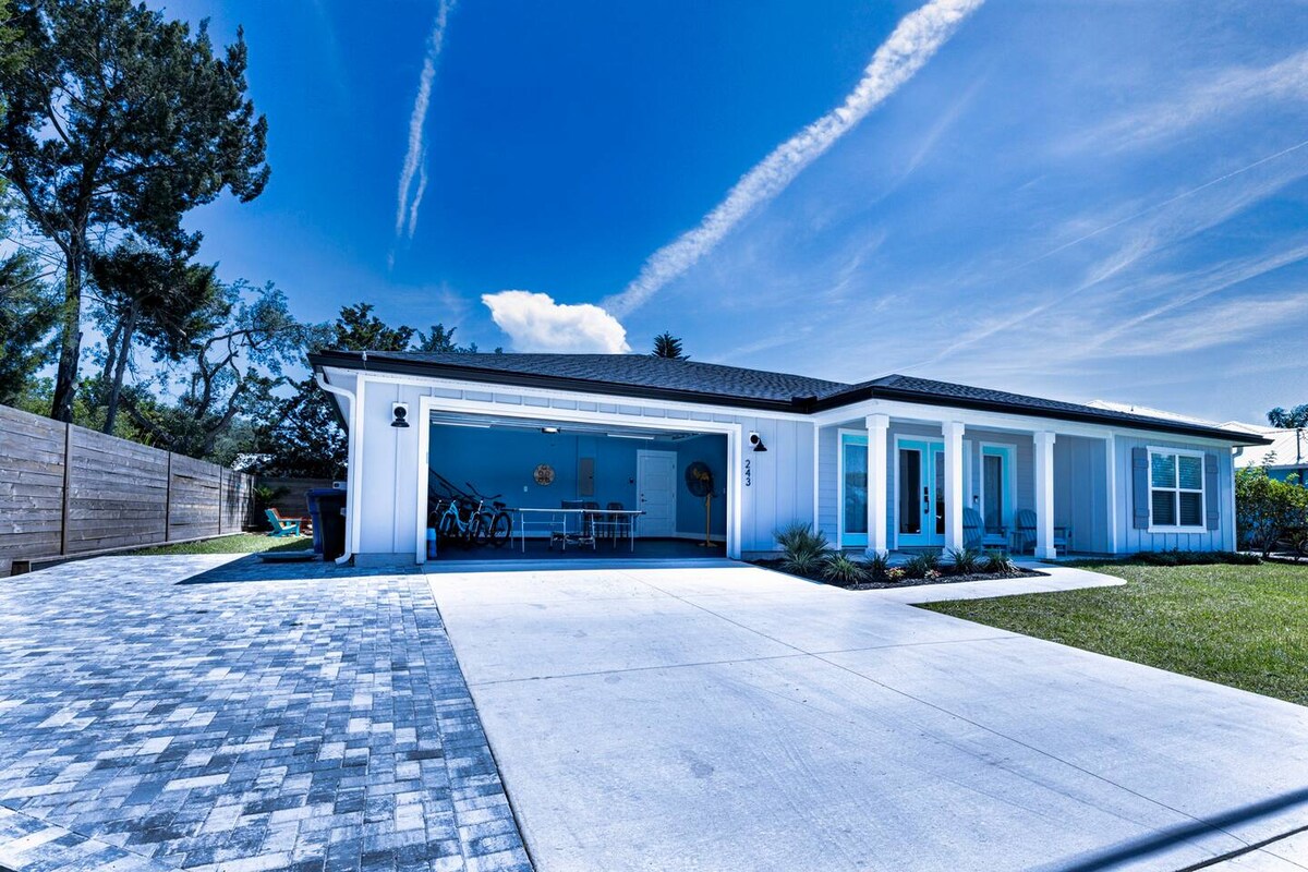 The exterior of the home is showcased with a paved driveway and green lawn. A spacious garage is visible, housing bicycles and outdoor furniture. The bright blue sky complements the light-colored house, enhancing the inviting atmosphere of the property.