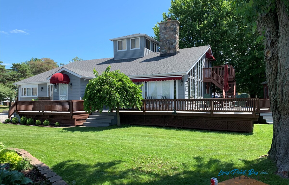 A spacious cottage with a gray exterior and large windows is surrounded by a well-maintained lawn. A multi-level deck with railing offers outdoor seating. A natural stone hearth feature is visible, and a red awning accents the entrance.