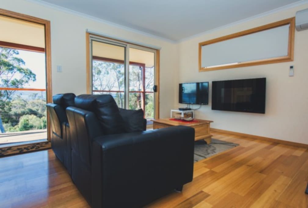 A cozy living area features a pair of black armchairs, positioned near large glass doors that allow natural light in. A flat-screen TV is mounted on the wall, and a wooden coffee table rests on the warm wooden floor, creating a comfortable space to relax.